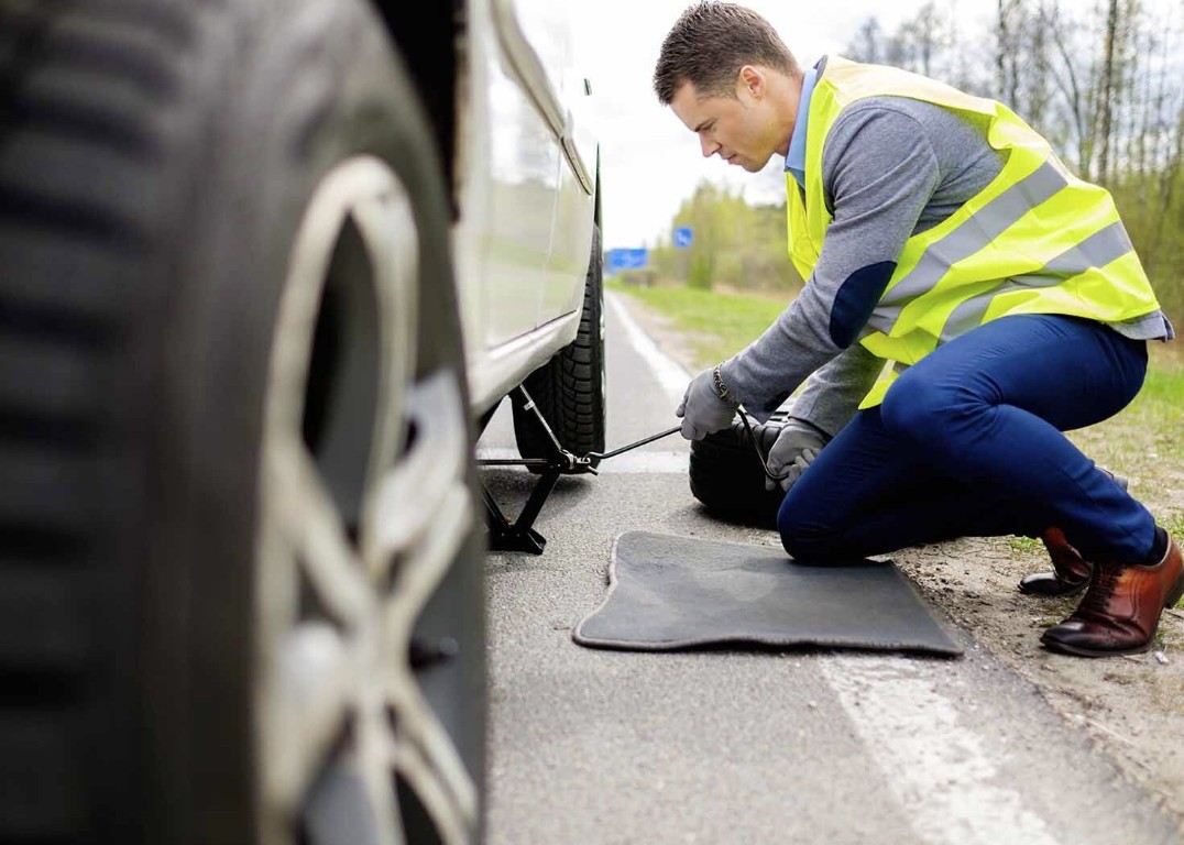 Roadside technician in a safety vest changing a flat tire on the road shoulder in North Babylon, NY